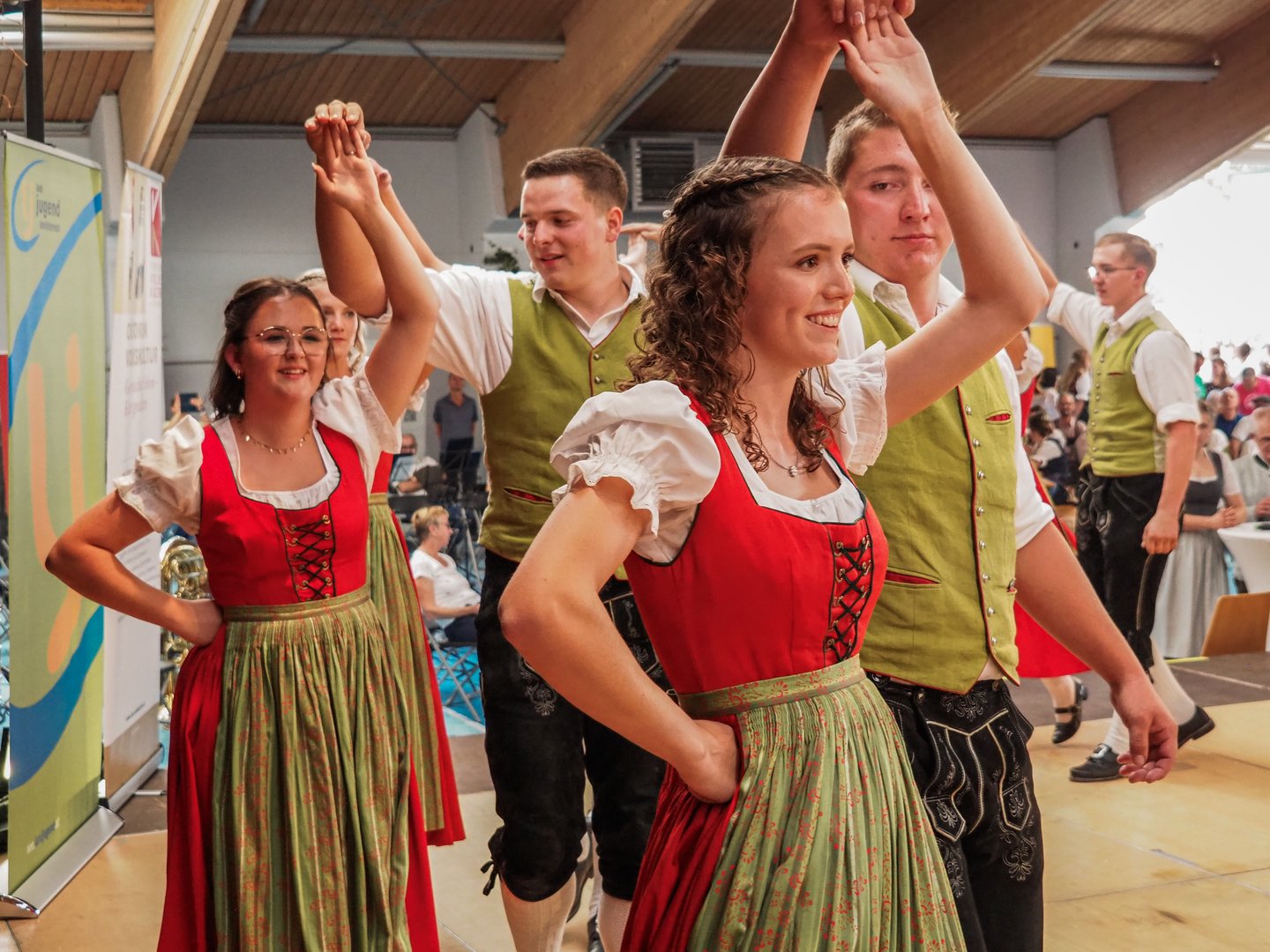 Trachtengruppe tanzt mit erhobenen Armen in Tracht, junge Frau im Vordergrund.